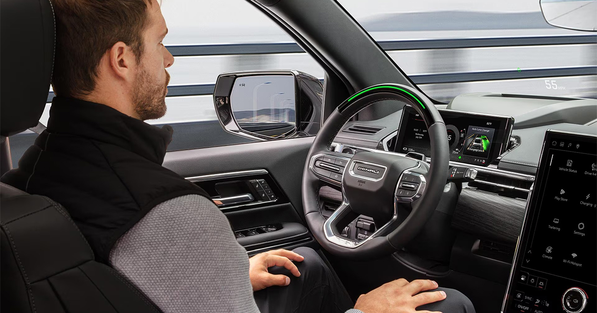 Driver sitting in a GMC Denali with hands off the wheel as the Super Cruise hands-free driving system operates, interior screens showing speed and lane guidance.