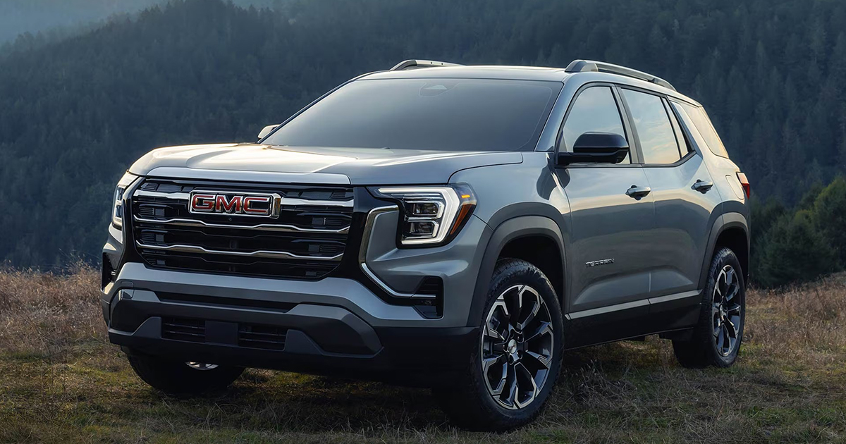Front view of a silver 2026 GMC Terrain SUV parked outdoors, showcasing its bold grille with a red GMC logo, modern LED headlights, black alloy wheels, and rugged styling against a backdrop of trees and mountains.