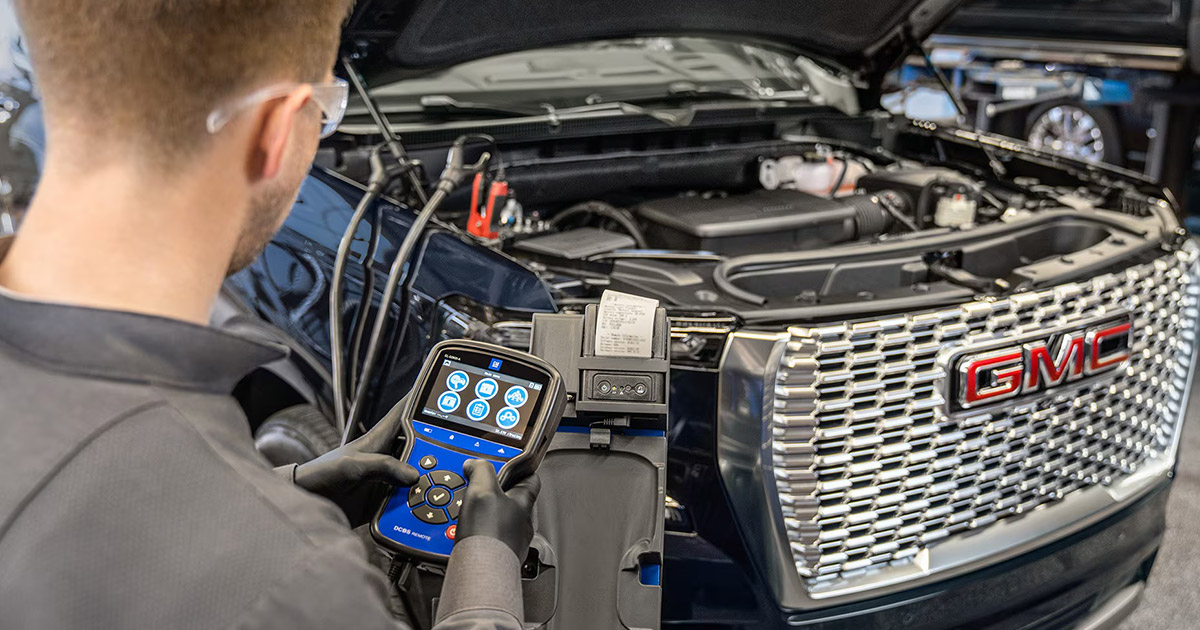 Technician using a diagnostic tool to test the battery and electrical system of a GMC vehicle with the hood open, focusing on the front grille with the red GMC logo and engine components visible.