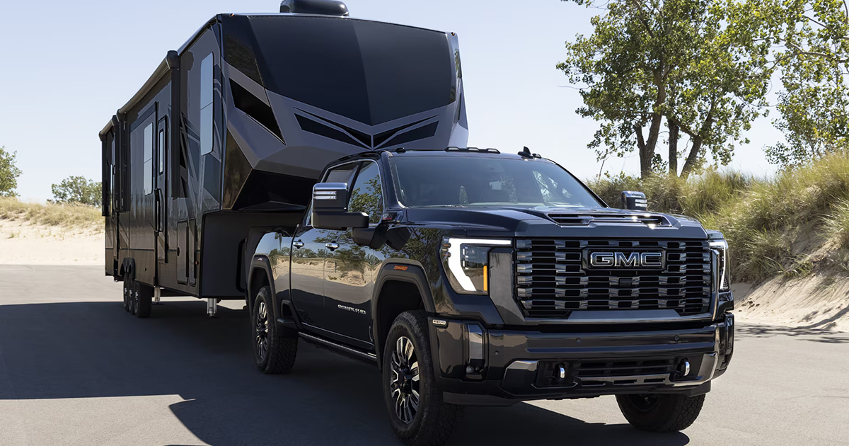 Black GMC Sierra HD pickup truck towing a large modern fifth-wheel trailer on a paved road, featuring a bold front grille with GMC logo, chrome accents, and rugged styling, with trees and sand dunes in the background.