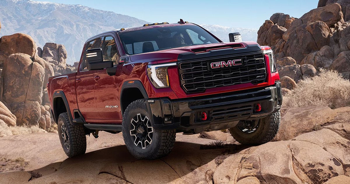 Red GMC Sierra AT4X HD pickup truck climbing over rugged rocks in a desert mountain landscape, highlighting its off-road suspension and bold front grille.