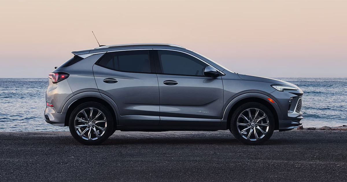 Side profile of a silver Buick Encore GX parked near the ocean at sunset, emphasizing its sleek body lines and modern SUV design.
