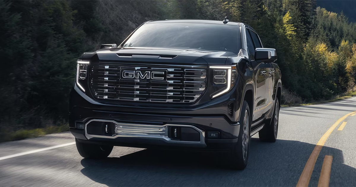 Black GMC Sierra pickup truck driving on a forested highway, showcasing its chrome grille and bold styling.