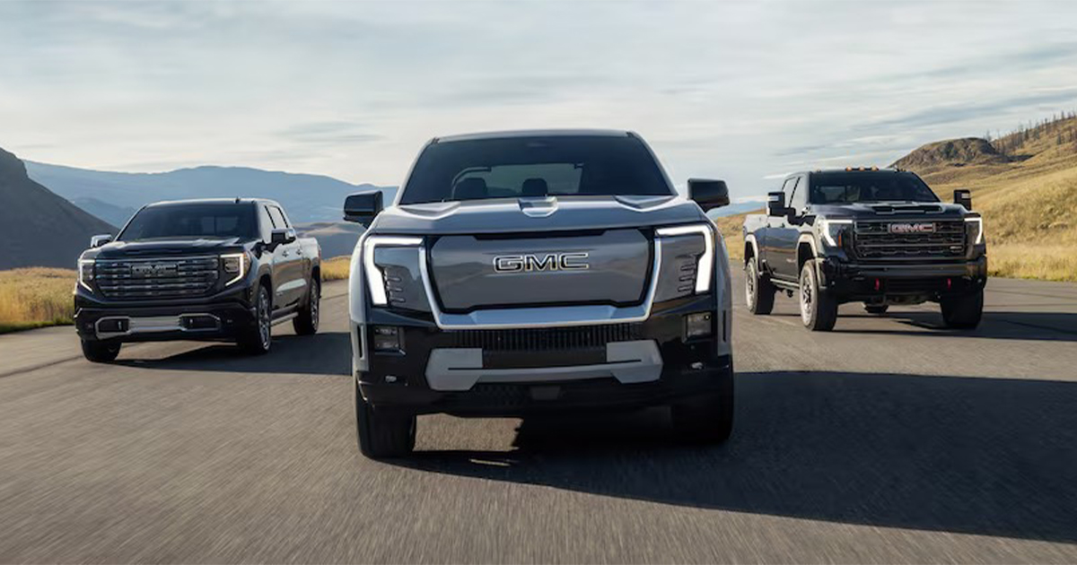 Front view of three GMC pickup trucks driving side by side on a highway, with the GMC Sierra EV in the center, flanked by two traditional GMC trucks in a scenic mountain landscape.