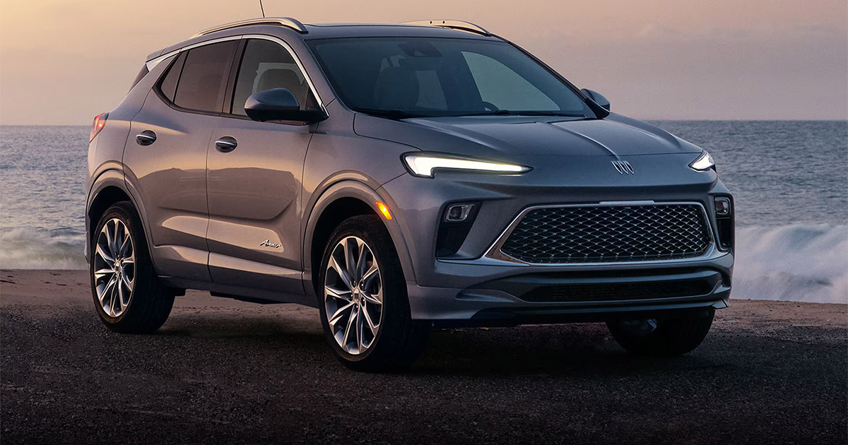 Silver Buick Encore GX parked near the ocean at dusk, with headlights on and modern grille design visible against the waves in the background.