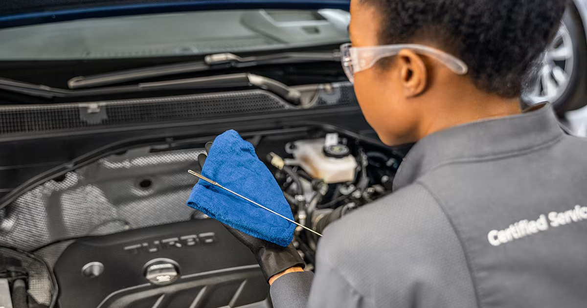 Certified Service technician wearing gloves and safety glasses inspects an engine oil dipstick while holding a blue cloth under the hood of a turbocharged vehicle.