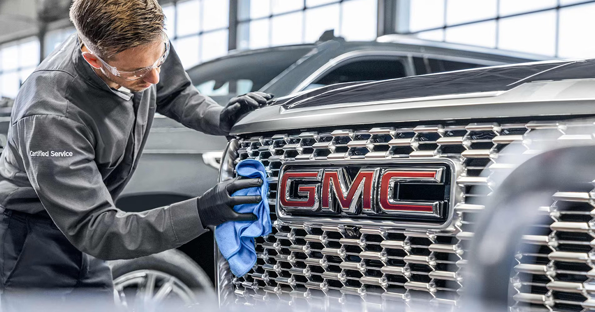 Certified Service technician wearing safety glasses wipes the chrome grille of a GMC vehicle with a blue cloth, highlighting the red GMC emblem.