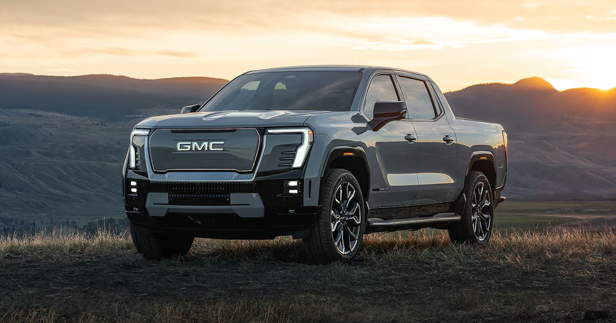 Front view of three GMC pickup trucks driving side by side on a highway, with the GMC Sierra EV in the center, flanked by two traditional GMC trucks in a scenic mountain landscape.