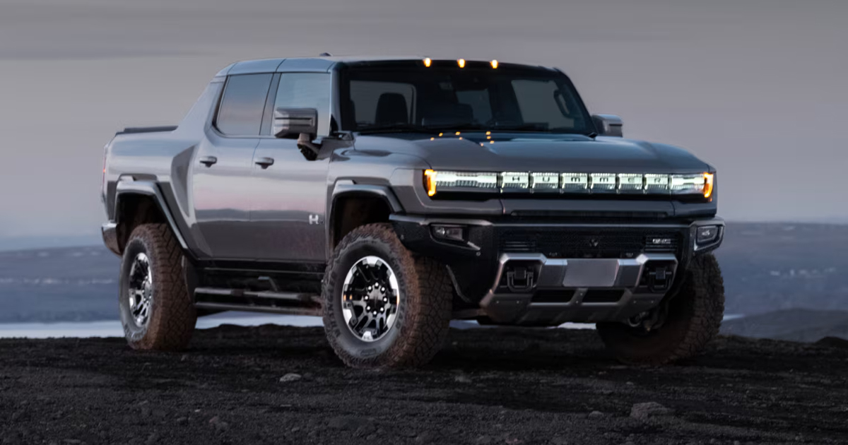 GMC Hummer EV pickup truck with rugged off-road tires and illuminated front light bar, parked on rocky terrain at dusk with water in the background.